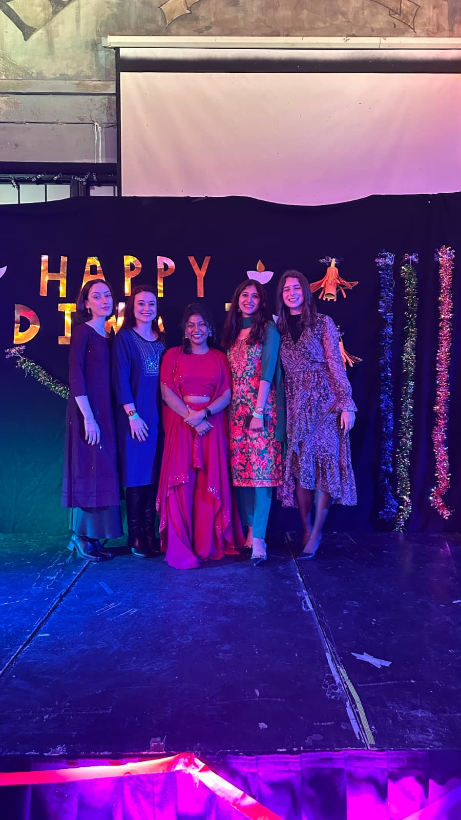 Five friends in traditional Indian clothing posing for a photo in front of a sign that reads "Happy Diwali" 